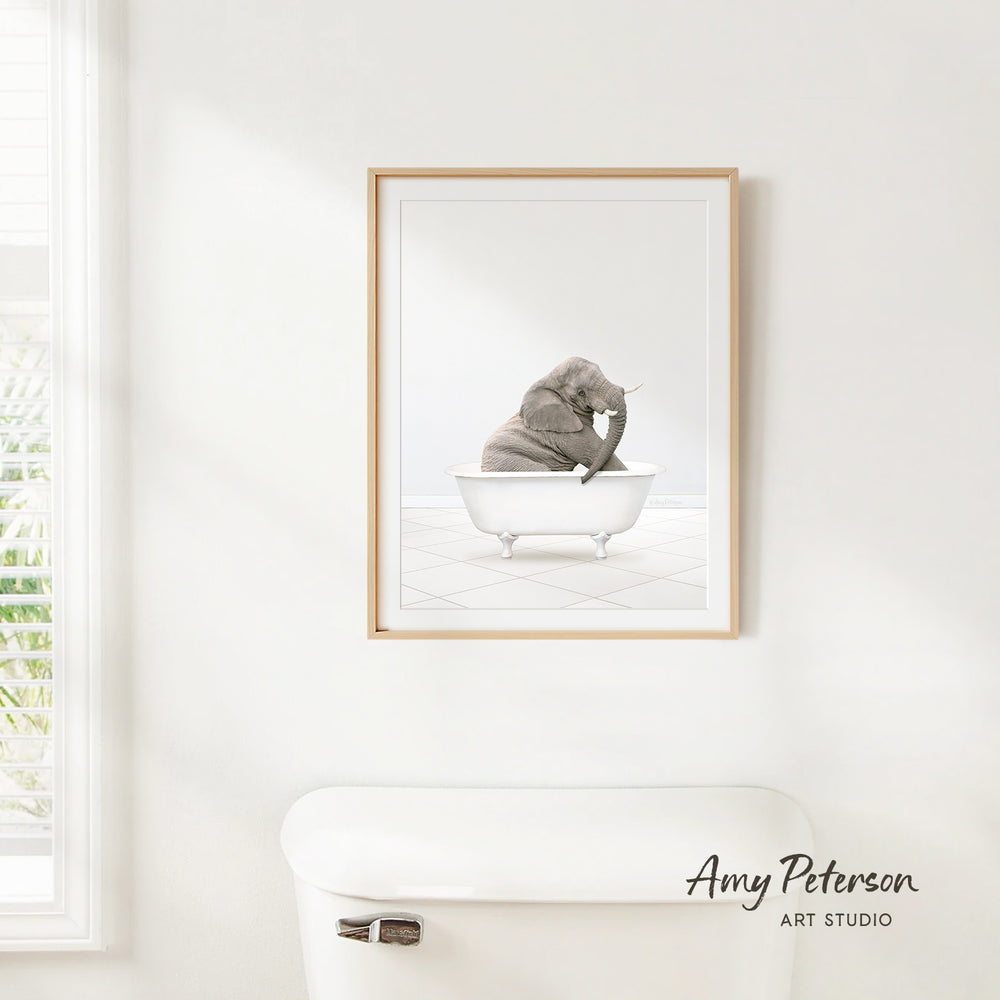 A framed image of a baby elephant sitting in a bathtub, with a window and a white toilet visible in the background.