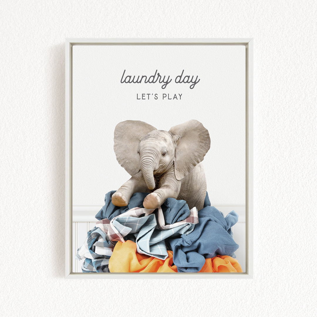 A framed image of a stuffed elephant sitting atop a pile of laundry, with the text "laundry day let's play" written above it.