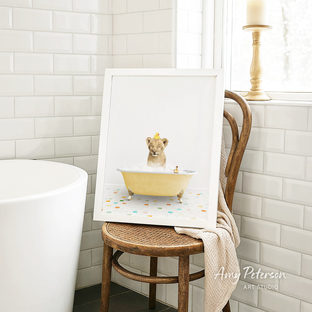A framed image of a lion sitting in a yellow bathtub, with a towel draped over the back of a wooden chair in a bathroom setting.