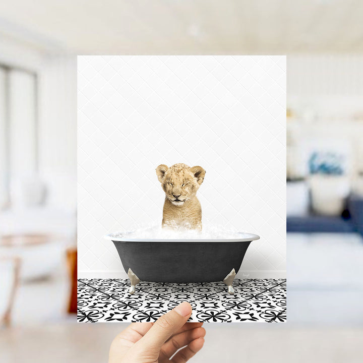 A hand holds up a framed image of a young lion cub sitting in a bathtub filled with water, surrounded by a black and white patterned floor.