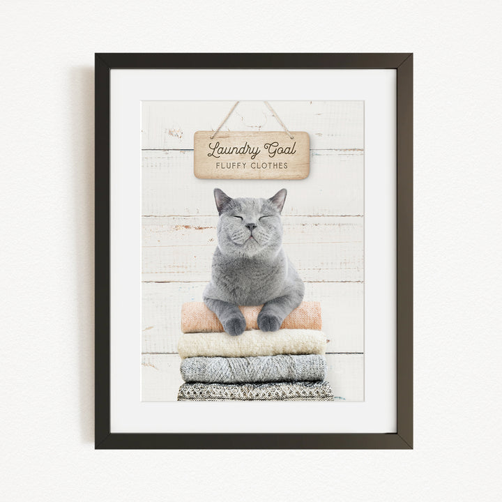 A framed image of a gray cat sitting on a stack of folded clothes, with a sign above that says "Laundry goal fluffy clothes".