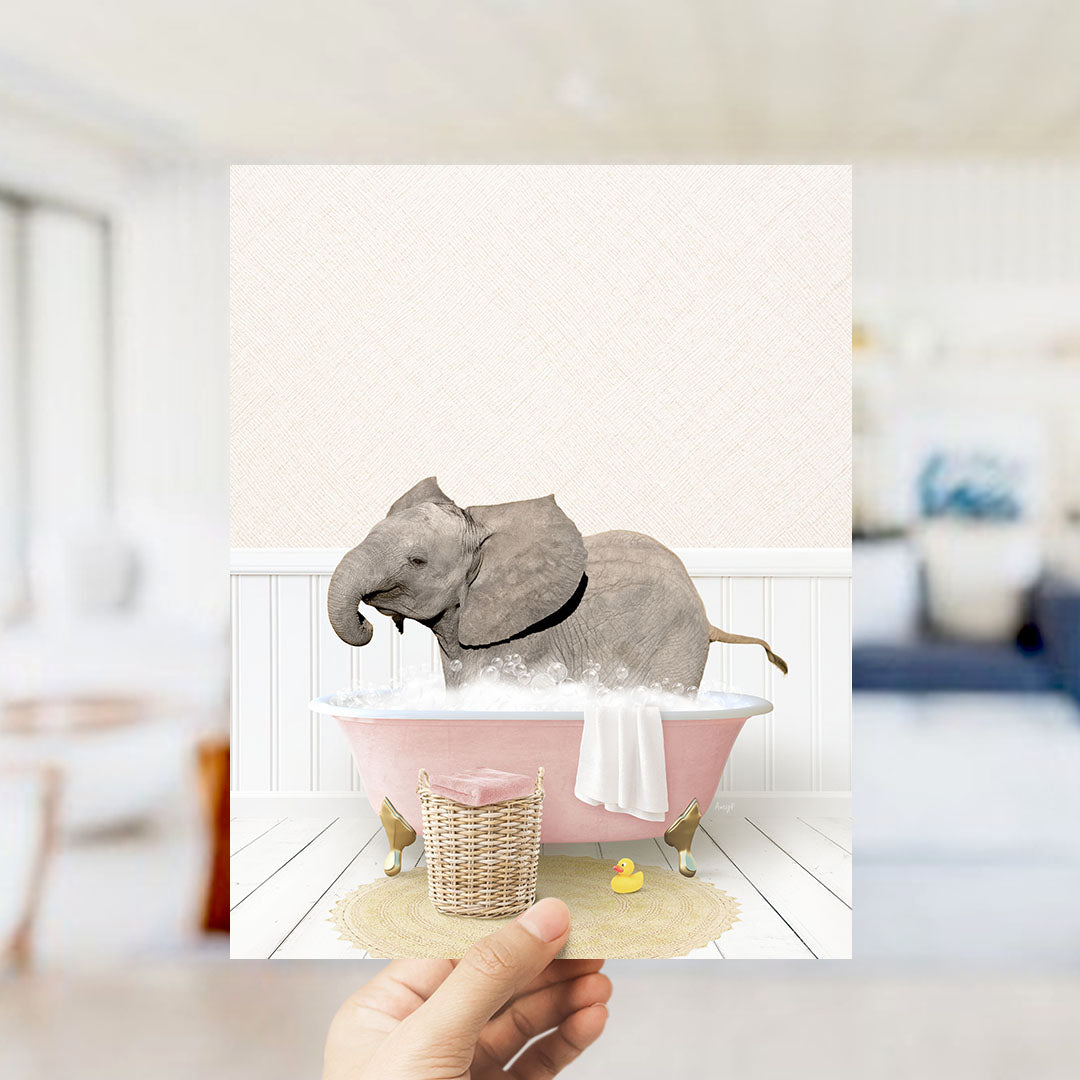 A hand holds up a framed image depicting a baby elephant sitting in a pink bathtub filled with bubbles, surrounded by a yellow rubber duck and a white towel.