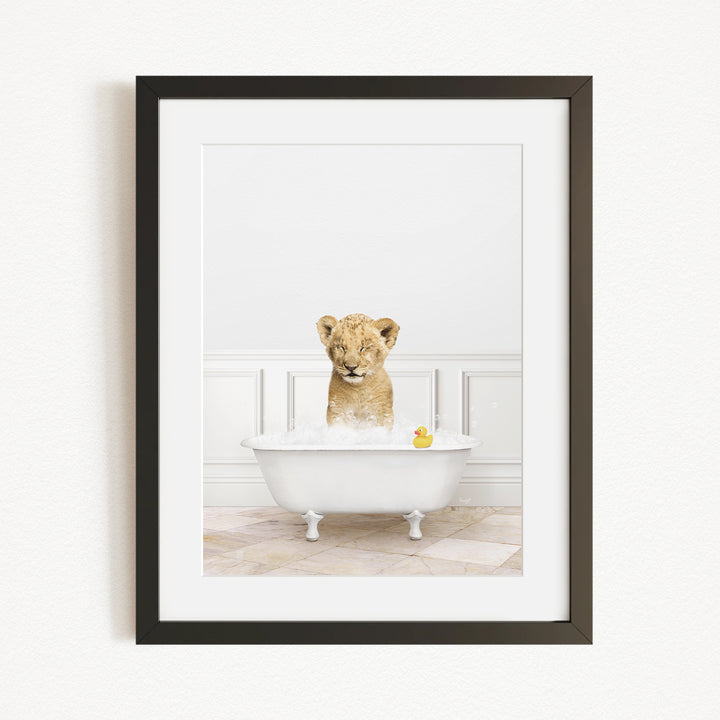 A young lion cub sitting in a white bathtub filled with water and bubbles, with a yellow rubber duck floating in the water.