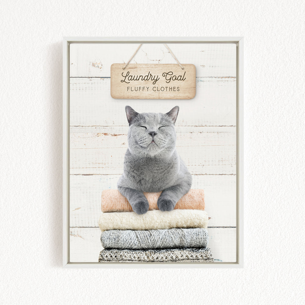 A gray cat sitting on a stack of clothes with a wooden sign above it that says "Laundry Goal Fluffy Clothes".