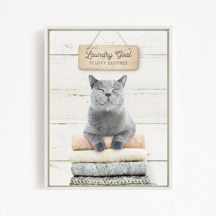 A gray cat sitting on a stack of clothes with a wooden sign above it that says "Laundry Goal Fluffy Clothes".