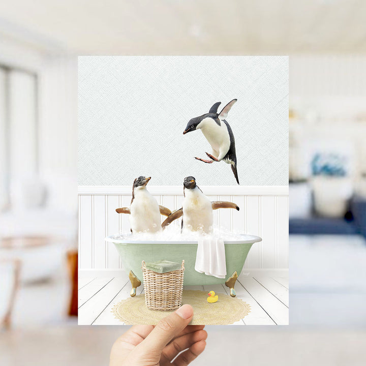 A hand holds up a framed image depicting two penguins sitting in a bathtub, with one penguin flying towards the other.