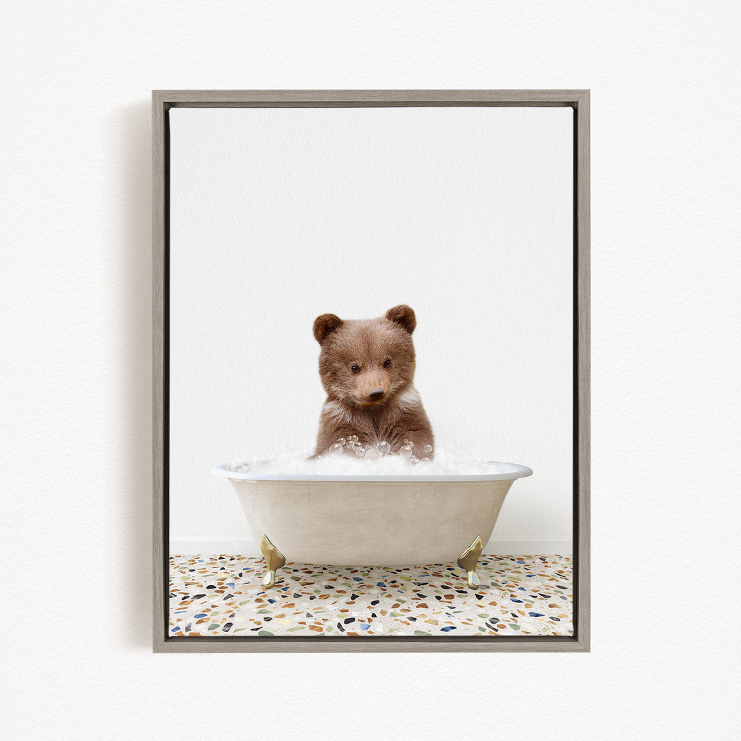 A cute brown bear cub sitting in a white bathtub filled with water, looking directly at the camera.