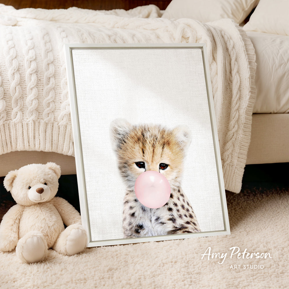 A framed print of a cheetah blowing a bubble with a pink bubble gum bubble is displayed on a bed, accompanied by a teddy bear.