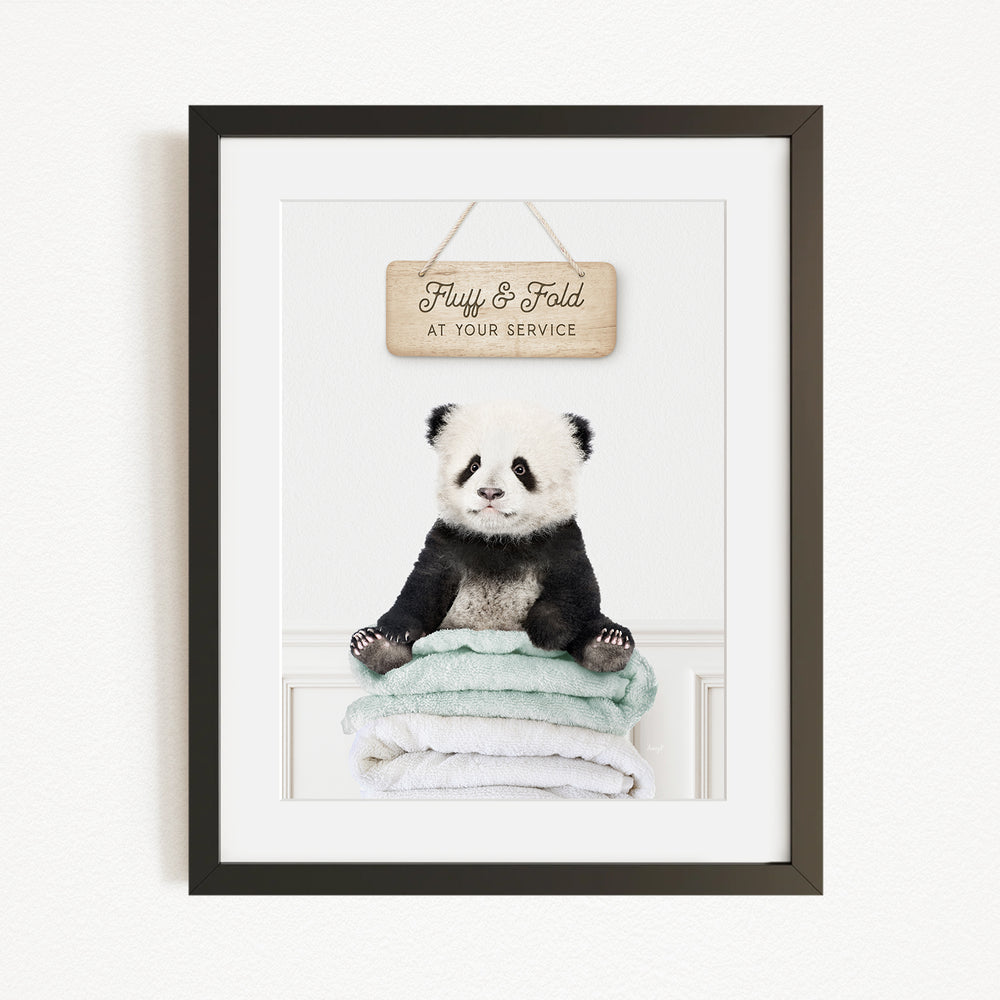 A panda bear sitting on a stack of towels, with a wooden sign above that says "Fluffy & Fold at your service".