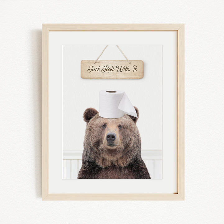 A framed image of a brown bear wearing a toilet paper hat, with a wooden sign above it that says "Just Roll With It".