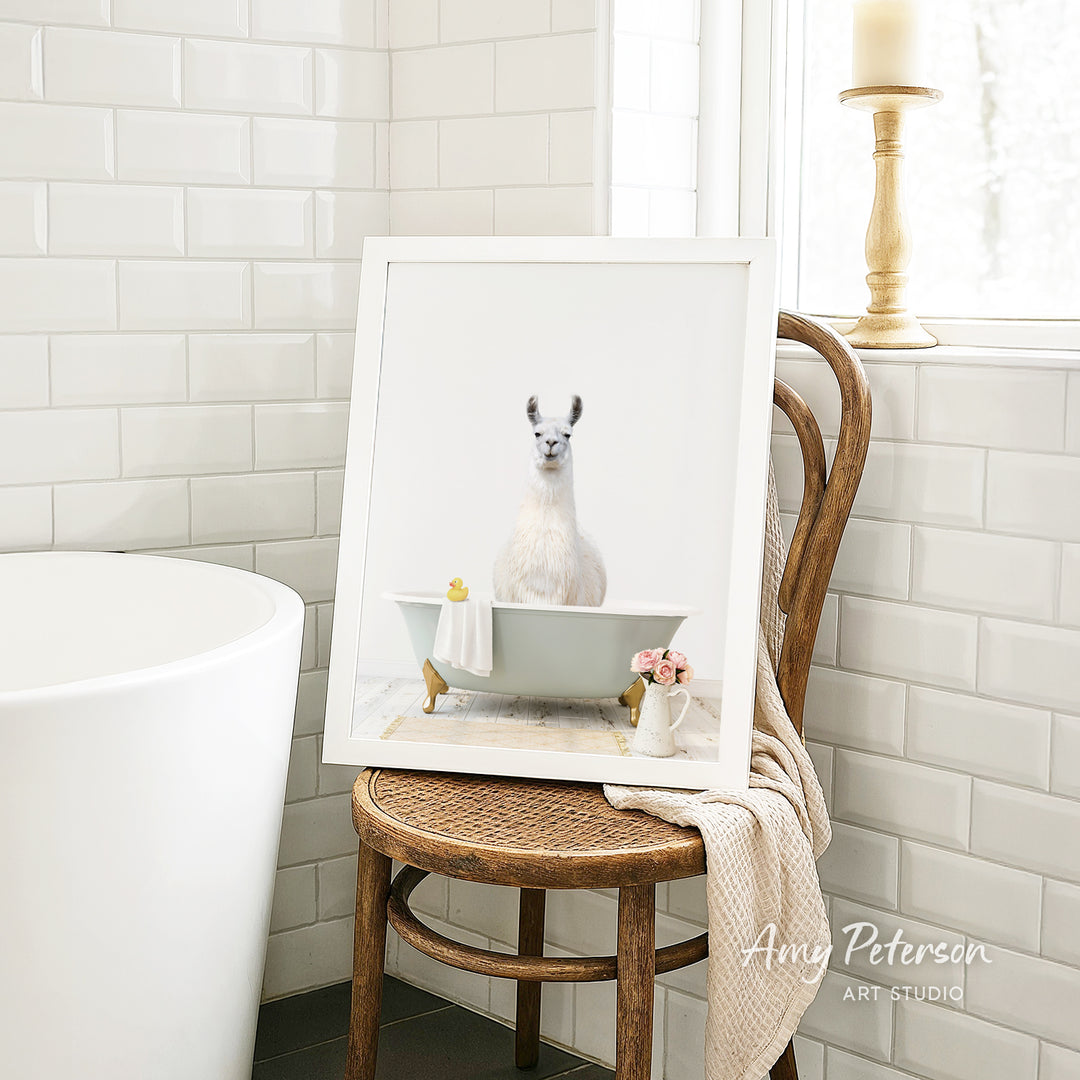A framed image of a white llama sitting in a bathtub, with a vase of flowers and a towel nearby, is displayed on a wooden chair in a bathroom setting.