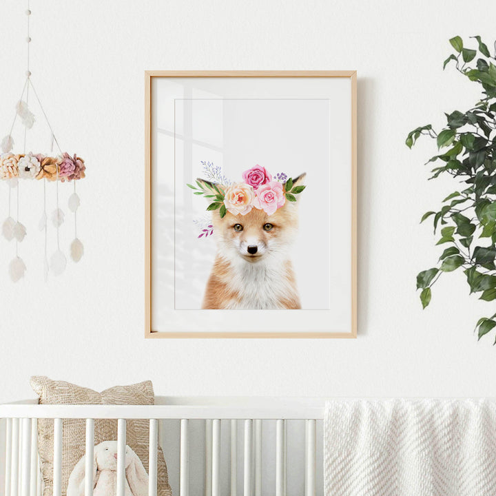 A framed print of a fox wearing a flower crown is displayed on a white wall, with a crib and a hanging mobile with flowers in the background.