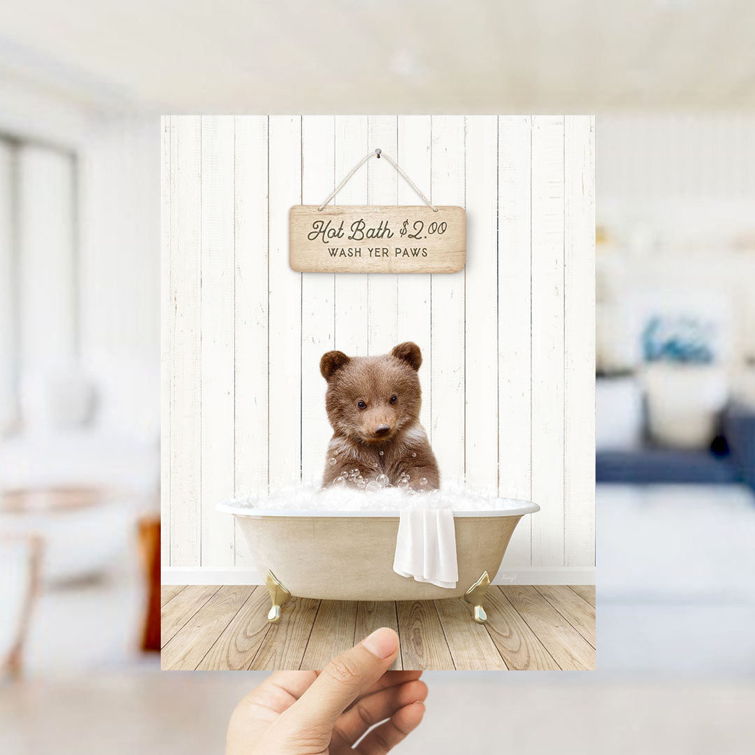 A hand holding a framed image of a brown bear sitting in a bathtub, with a sign above it that reads "Hot Bath $2.00 WASH YER Paws".