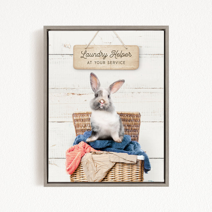 A gray and white rabbit sitting in a wicker basket filled with clothes, with a wooden sign above that reads "Laundry Helper at your service".