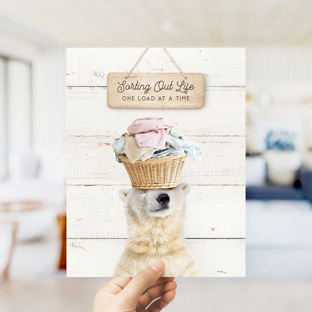 A hand holding a card with a polar bear standing on its head, wearing a basket of laundry, with a wooden sign above that says "Sorting Out Life One Load at a Time".