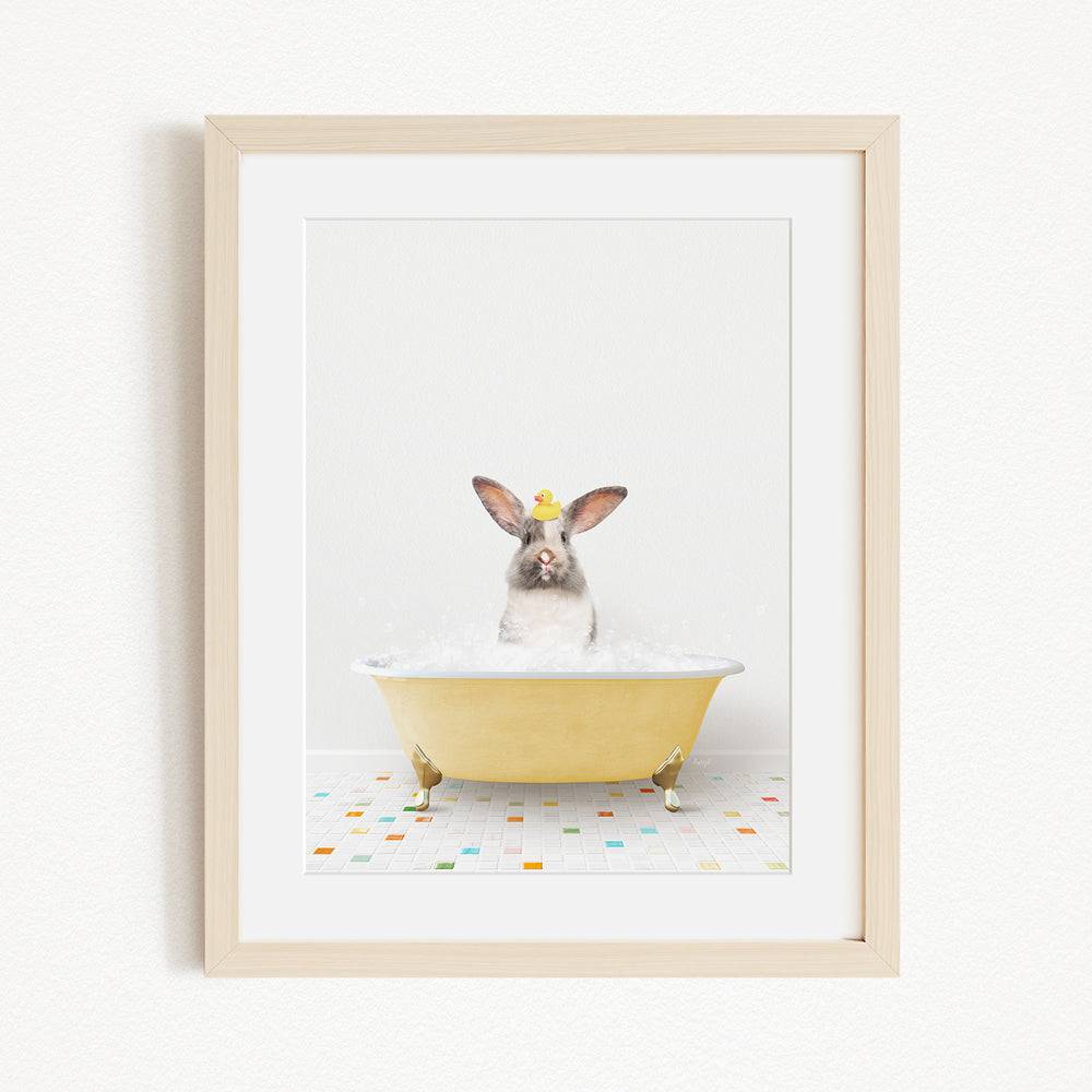 A gray rabbit wearing a yellow hat sits in a yellow bathtub filled with bubbles, surrounded by a colorful tiled floor.