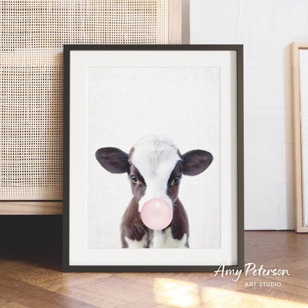 A framed image of a cow blowing a bubble with a pink bubble gum bubble.