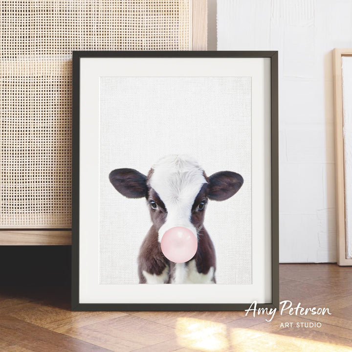 A framed image of a cow blowing a bubble with a pink bubble gum bubble.