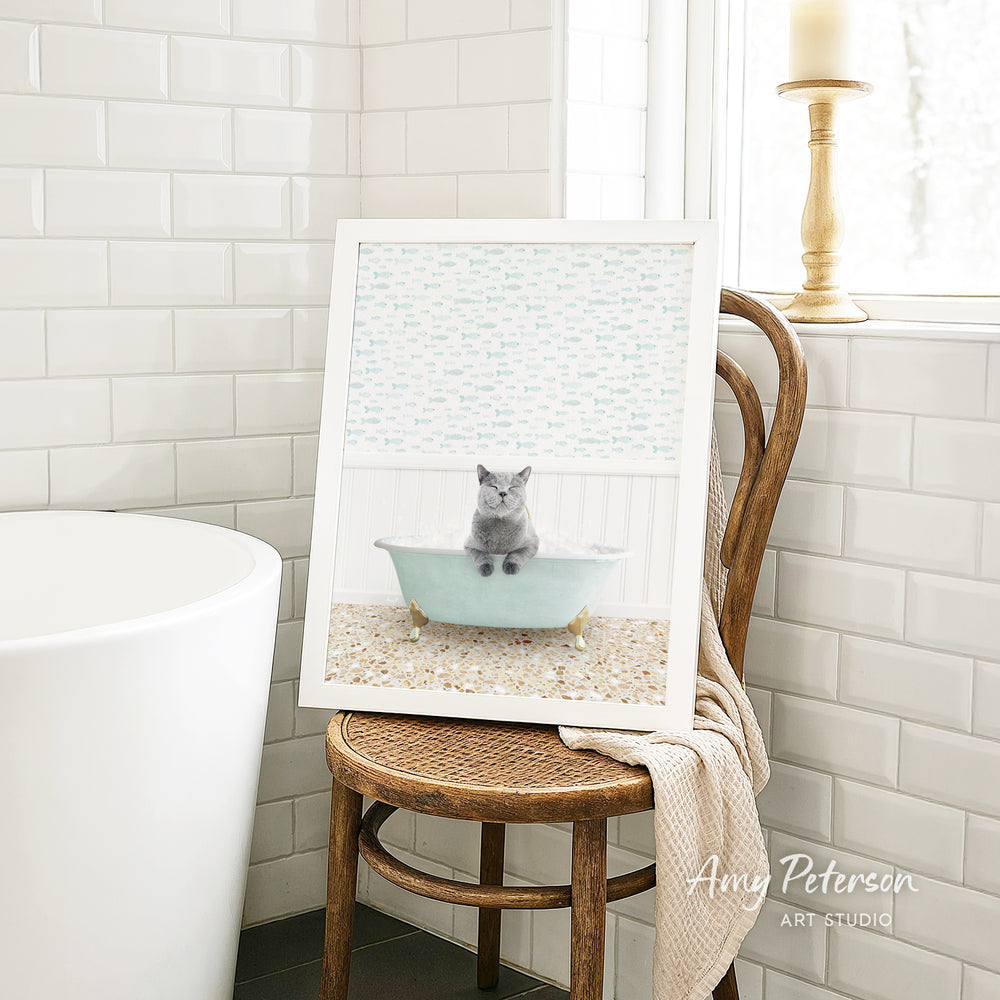 A framed image of a gray cat sitting in a bathtub, with a wooden chair and a white tiled wall in the background.