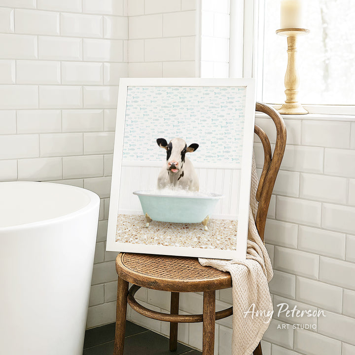 A framed image of a cow sitting in a bathtub is displayed on a wooden chair in a bathroom setting.