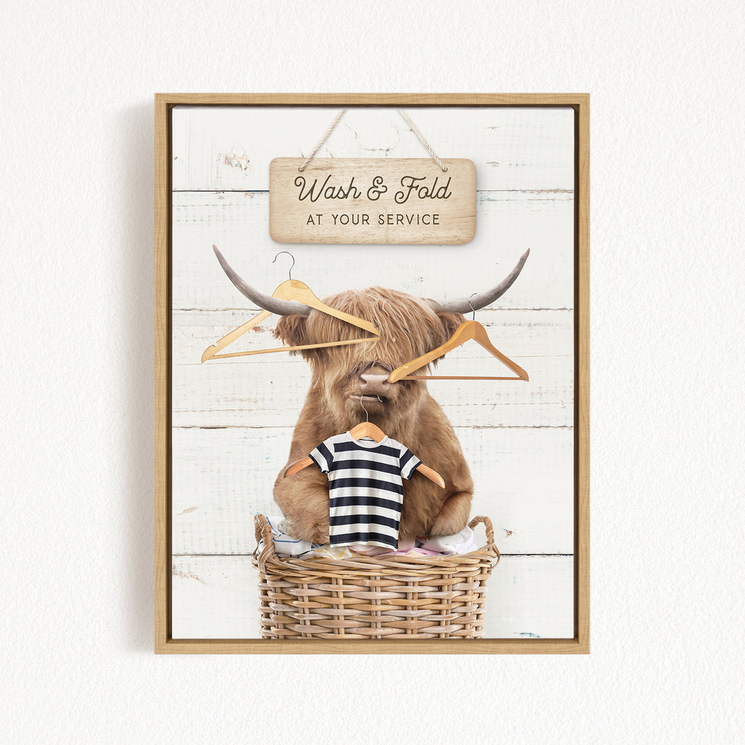 A framed image depicting a large, furry animal, possibly a yak or a yak dog, sitting in a wicker basket filled with clothes and hangers, with a sign above that reads "Wash & Fold at your Service".
