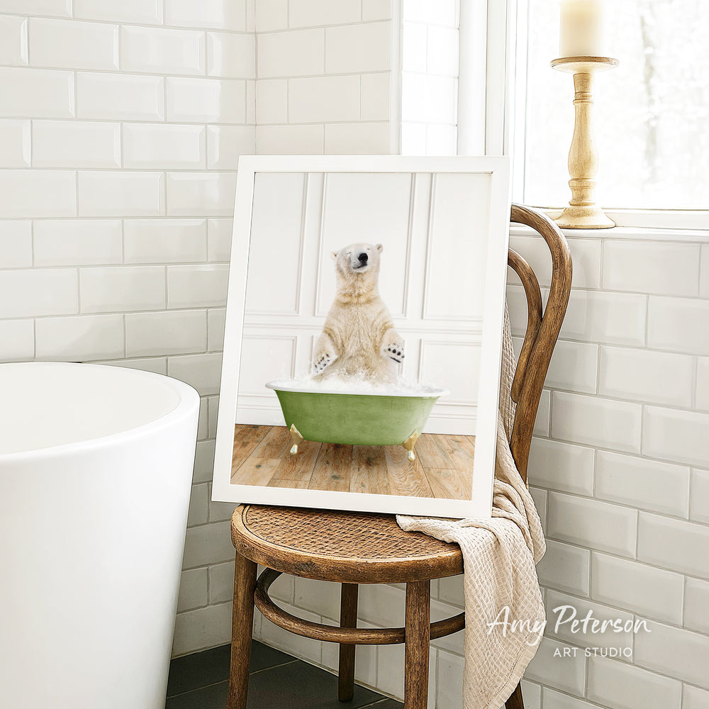 A framed image of a polar bear sitting in a green bathtub, with a towel draped over the back of a wooden chair in a bathroom setting.