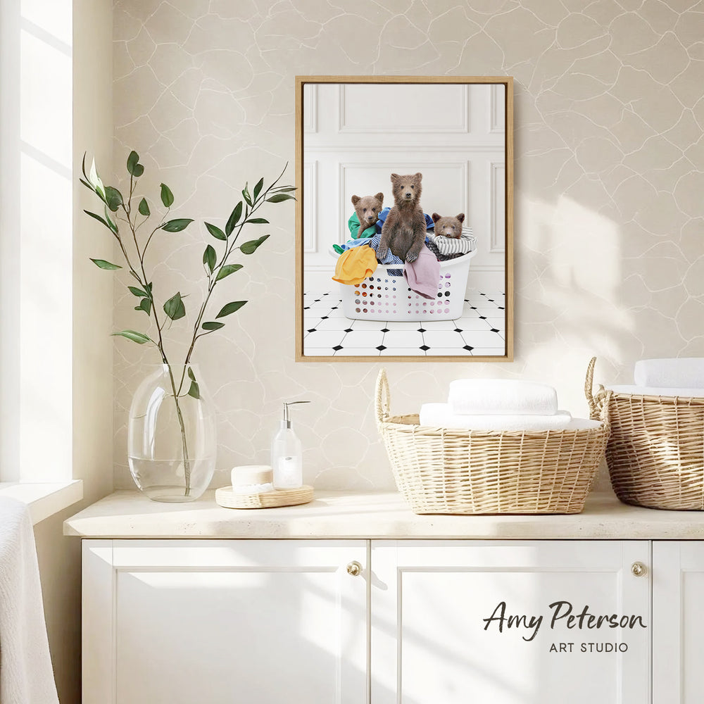 a framed photograph of three bears sitting in a laundry basket, with a vase of leaves and a bottle of soap on a white countertop.