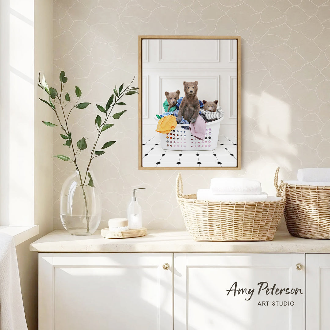 a framed photograph of three bears sitting in a laundry basket, with a vase of leaves and a bottle of soap on a white countertop.