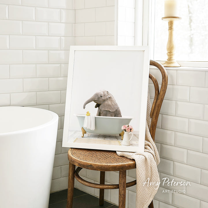A framed image of an elephant sitting in a bathtub, with a towel and flowers nearby, is displayed on a wooden chair in a bathroom setting.