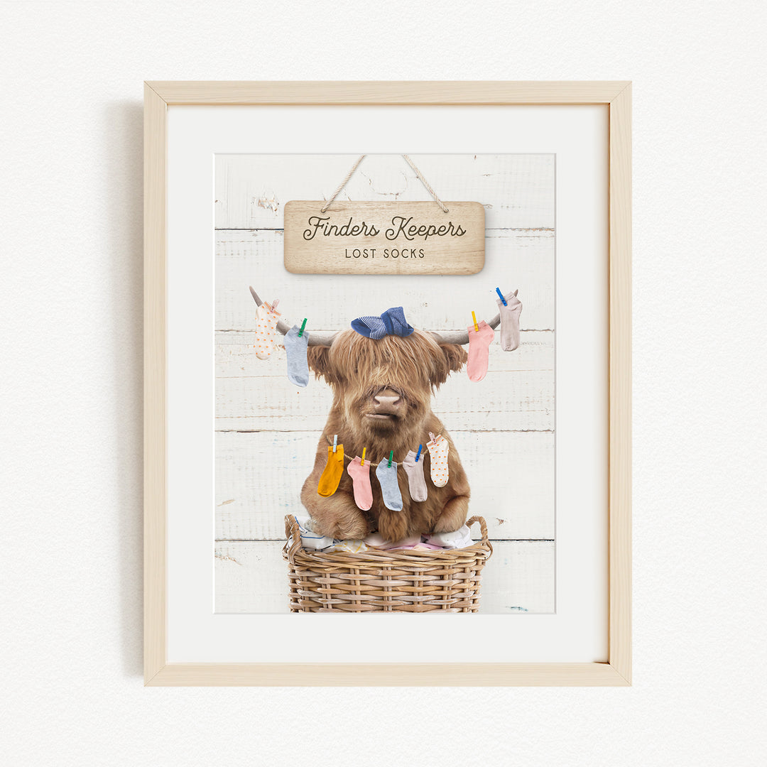 A framed image depicting a brown cow sitting in a wicker basket, with clothes hanging from a clothesline above it.