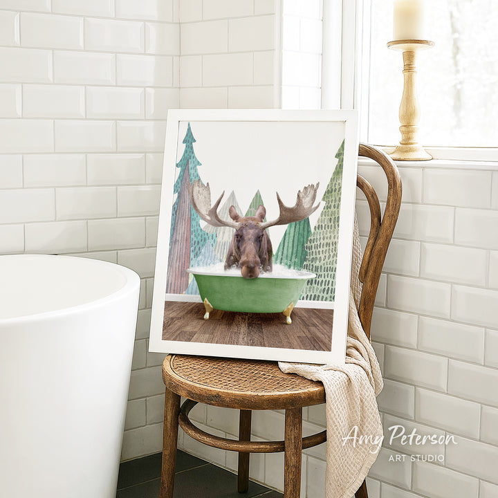 A framed image of a moose sitting in a green bathtub, surrounded by Christmas trees, is displayed on a wooden chair in a bathroom setting.