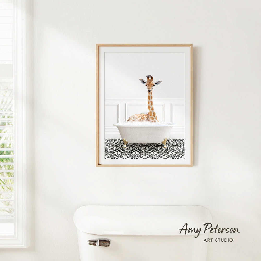 A giraffe sitting in a white bathtub, with a patterned tile floor beneath it, is the central focus of the framed artwork displayed on the wall.