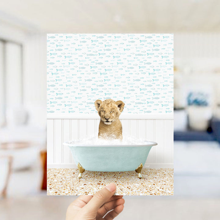 A hand holds up a collage of images depicting a young lion cub sitting in a bathtub filled with water, surrounded by a pattern of fish.