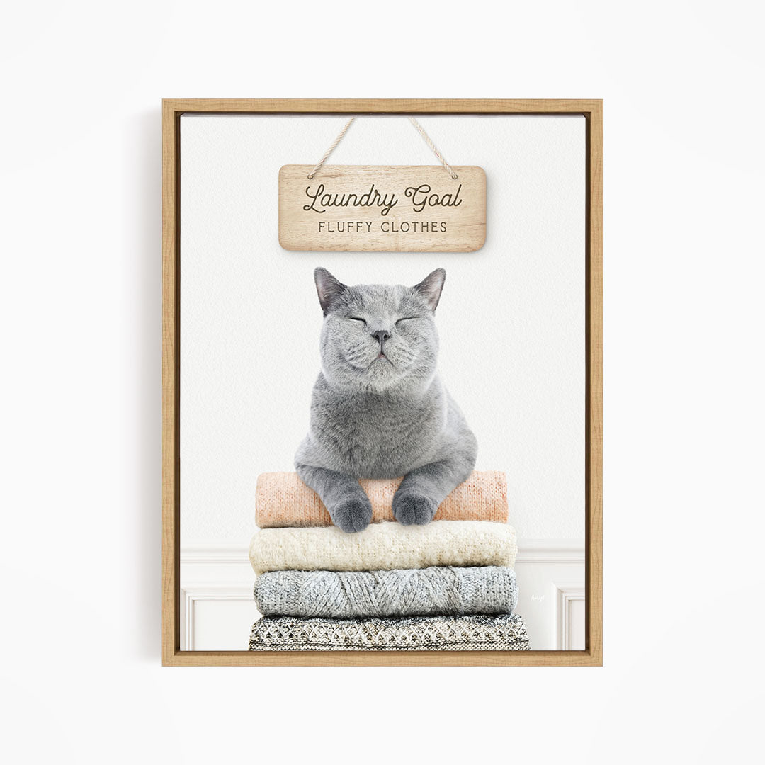 A gray cat sitting on a stack of folded clothes, with a wooden sign above that says "Laundry Goal Fluffy Clothes".