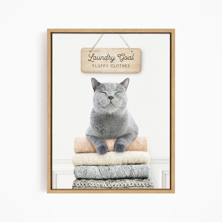 A gray cat sitting on a stack of folded clothes, with a wooden sign above that says "Laundry Goal Fluffy Clothes".