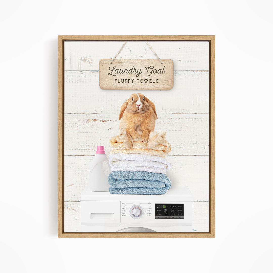 A framed image depicting a rabbit sitting atop a stack of fluffy towels, with a sign above that reads "Laundry Goal Fluffy Towels".