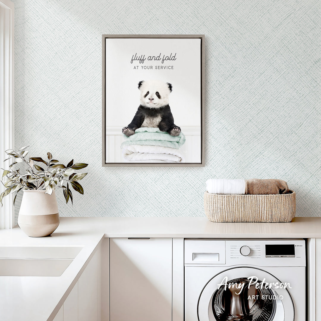 a framed poster of a panda bear sitting on a stack of towels, hanging on a wall above a white countertop with a basket and a plant.