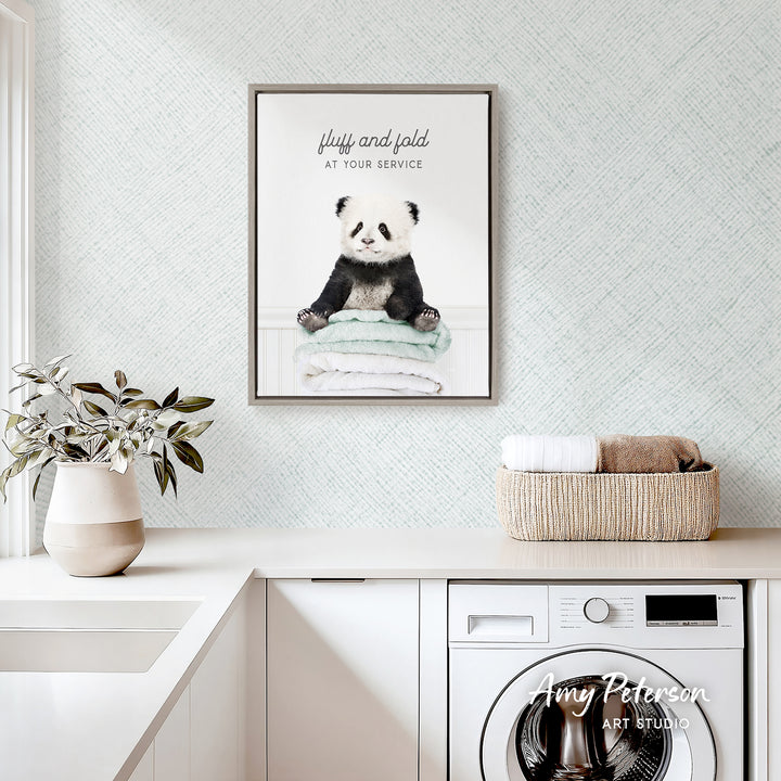 a framed poster of a panda bear sitting on a stack of towels, hanging on a wall above a white countertop with a basket and a plant.