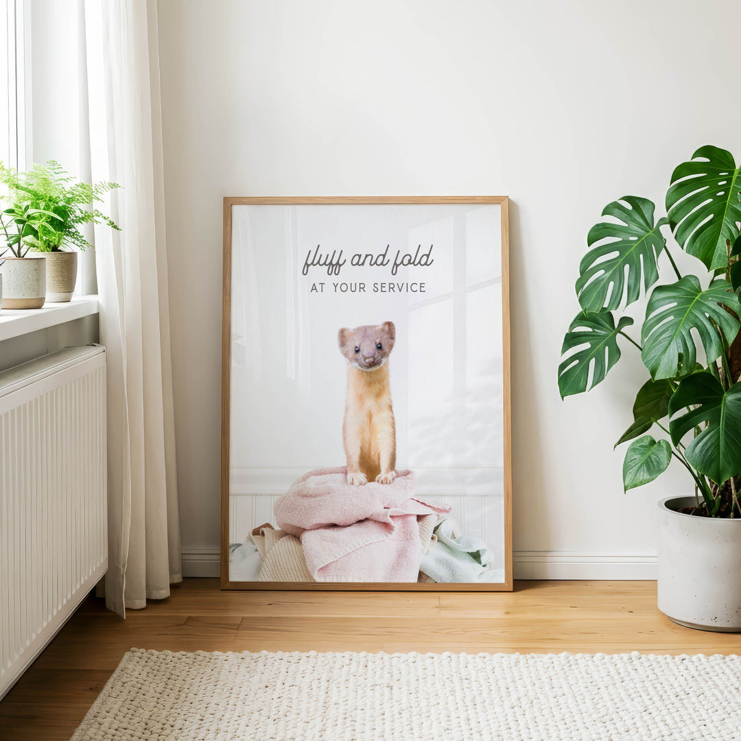 A framed poster with a cute cat sitting on a pink blanket, placed on a wooden floor next to a potted plant.