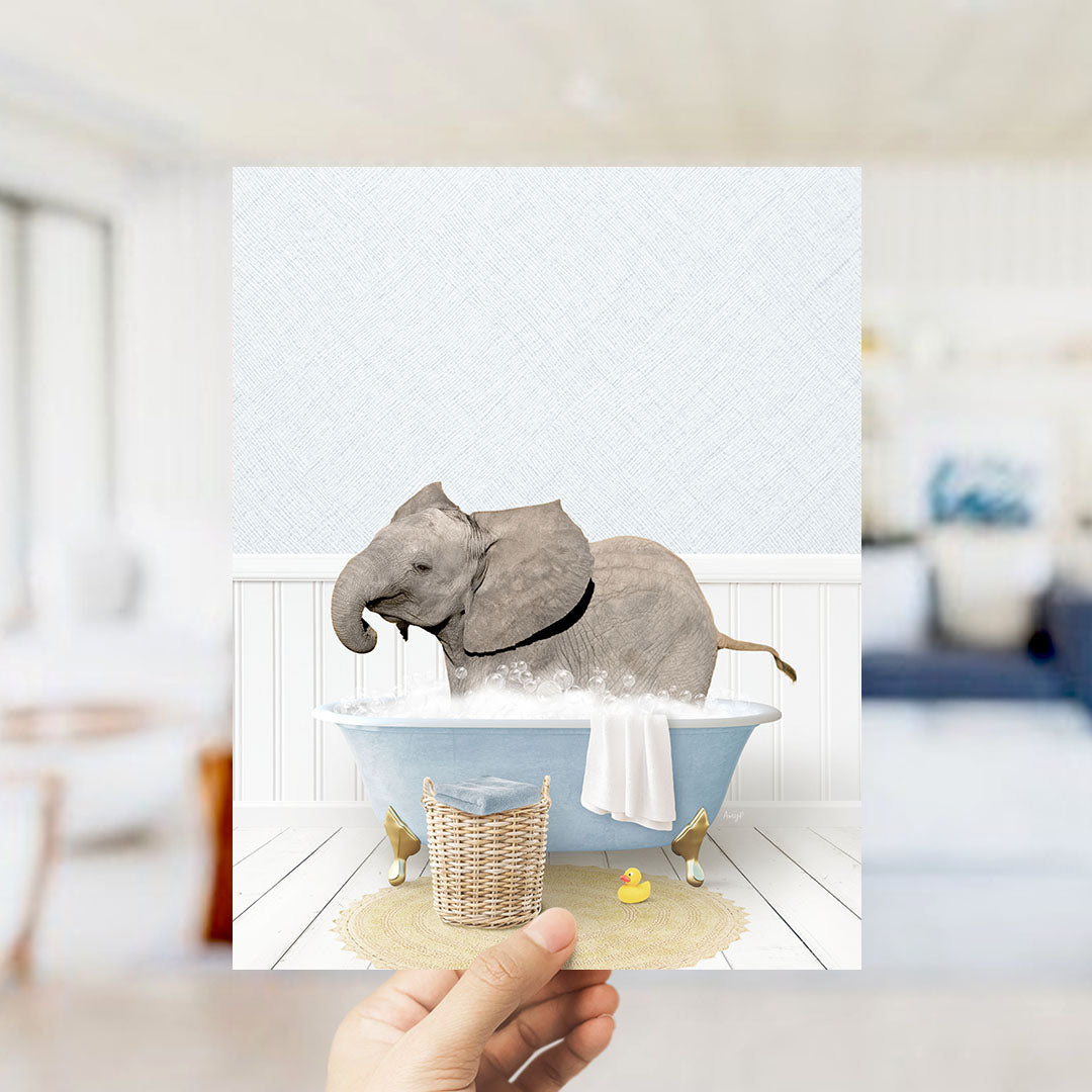 A hand holds up a framed image depicting a baby elephant enjoying a bath in a blue tub, surrounded by a yellow rubber duck and a white towel.
