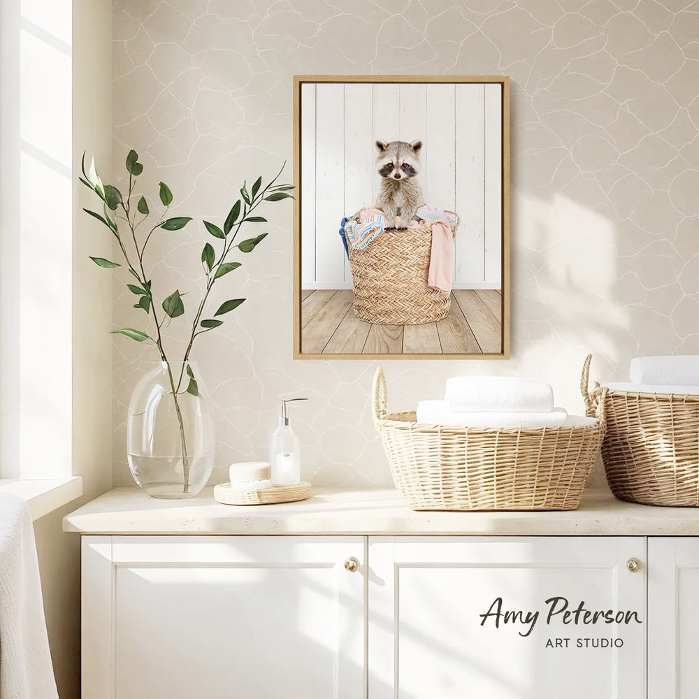 A framed photograph of a raccoon sitting in a woven basket is displayed on a wall above a white cabinet with baskets and a plant.