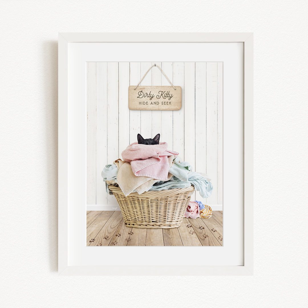 A framed image of a cat sitting in a basket filled with laundry, with a sign above the basket that reads "Dirty Kitty HIDE AND SEEE".