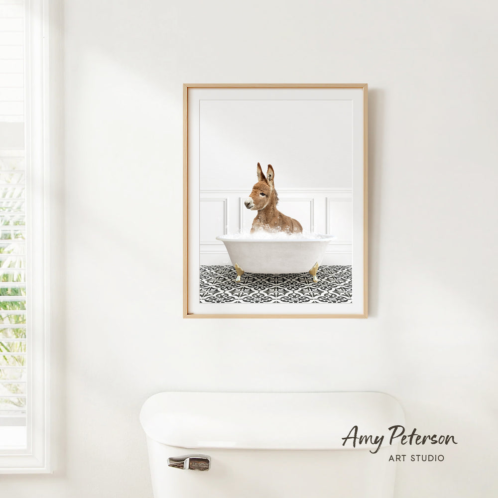 A framed image of a donkey sitting in a bathtub, with a patterned tile floor beneath it, is hanging on a white wall.