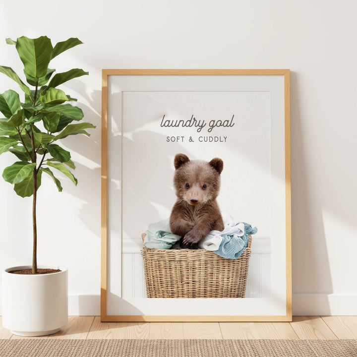 A framed image of a brown bear sitting in a wicker basket filled with clothes, with the text "laundry goal soft & cuddly" written above it.