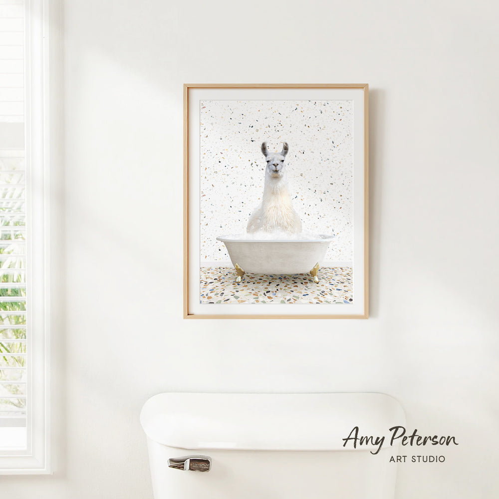 A framed image of a white llama sitting in a bathtub, surrounded by confetti, is hanging on a white wall above a white toilet.
