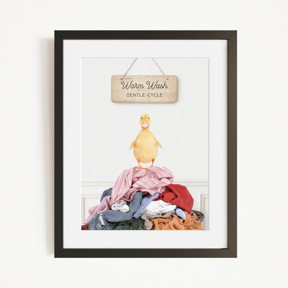 A framed image of a yellow duck standing on a pile of clothes, with a wooden sign above that reads "Warm Wash Gentle Cycle".