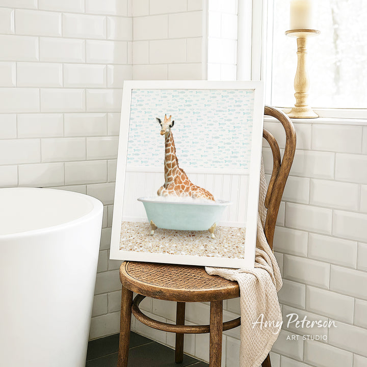 A framed image of a giraffe sitting in a bathtub is displayed on a wooden chair in a bathroom setting.