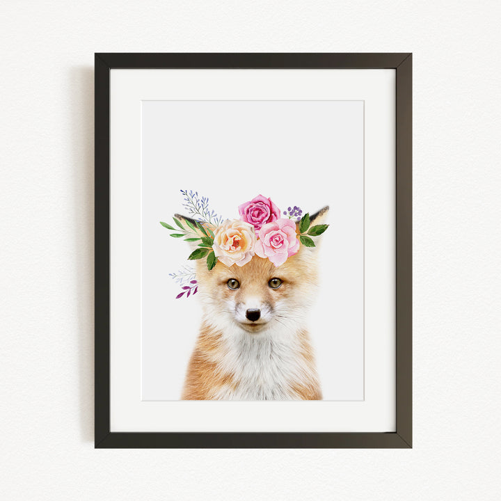 A framed print of a fox wearing a floral crown of pink and white roses and greenery.
