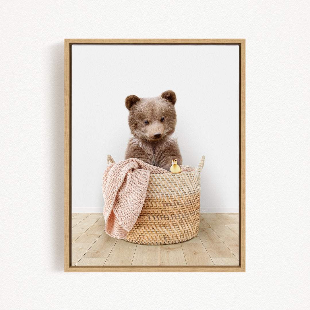A brown bear sitting in a woven basket, with a pink blanket and a small yellow toy duck nearby.
