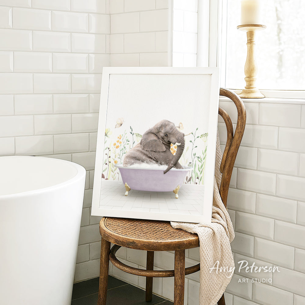 A framed illustration of a gray elephant sitting in a purple bathtub, surrounded by flowers, is displayed on a wooden chair in a bathroom setting.